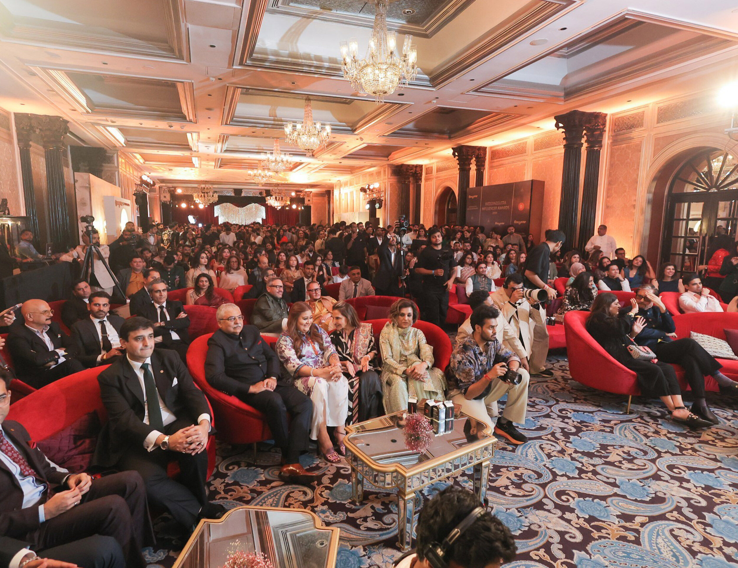 Grand indoor wedding celebration with elegantly dressed guests seated on red couches and a large crowd gathered under chandeliers in a luxurious hall.
